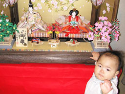 焼桐枠の台と桜柄の屏風が華やかな金彩衣装のお雛様を引き立てています。雛人形の前で赤ちゃんがこちらを向いて手を伸ばしています。