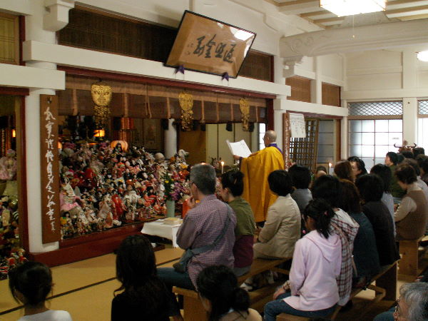 多聞院で開催される広島お人形供養会で住職が読経するワンシーン。参列者たちと山積みされた供養の人形。