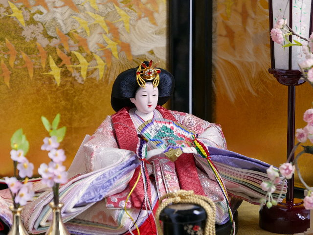 ほのかな赤と紫を組み合わせた優雅な光沢衣装の雛人形を鶴の金屏風と黒塗り台に並べる収納飾り