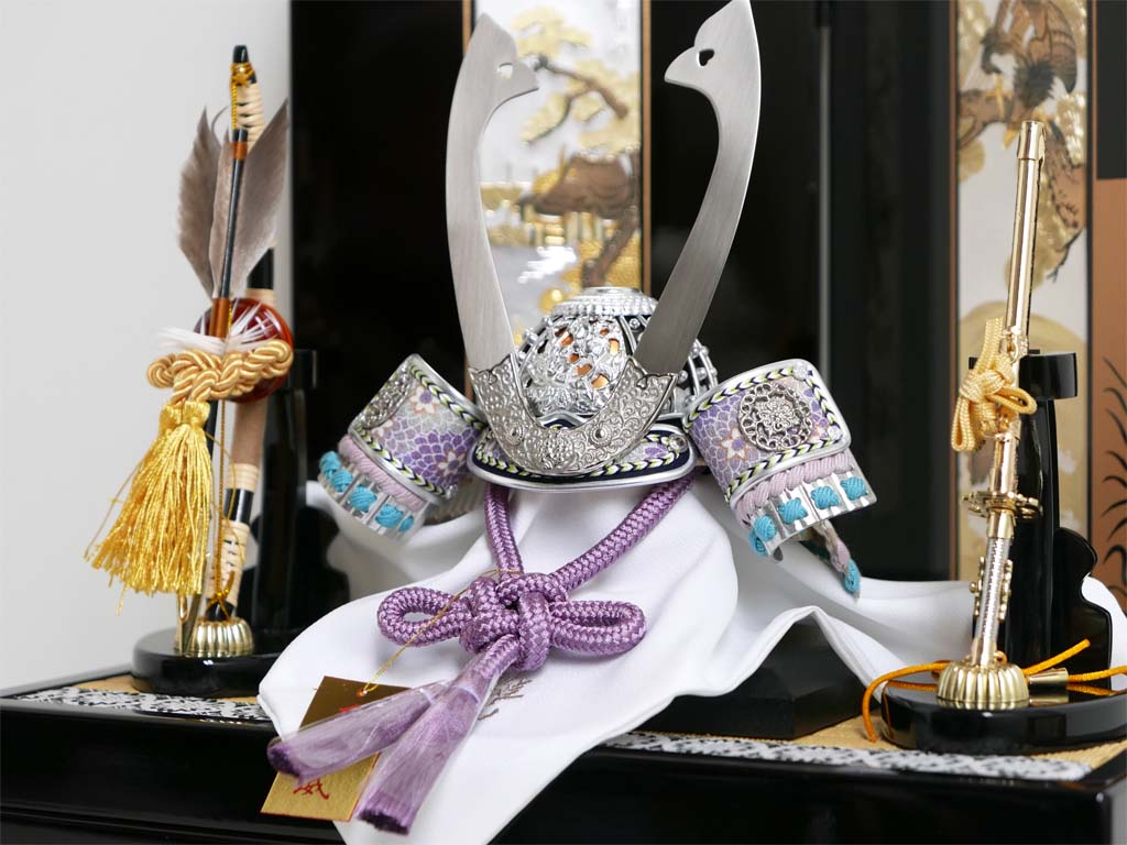 淡い色合いの威しを使い銀金物で統一した兜を、勇ましい松鷹がデザインされた彫金屏風に並べコンパクトな収納兜飾りにした五月人形です。