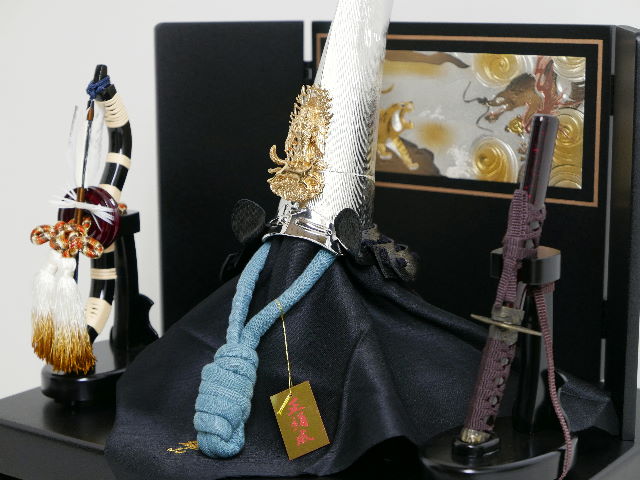 上杉謙信公仕様の銀烏帽子形兜を三曲の彫金龍虎屏風と一本矢弓太刀で黒塗りの収納台に並べた五月人形です。